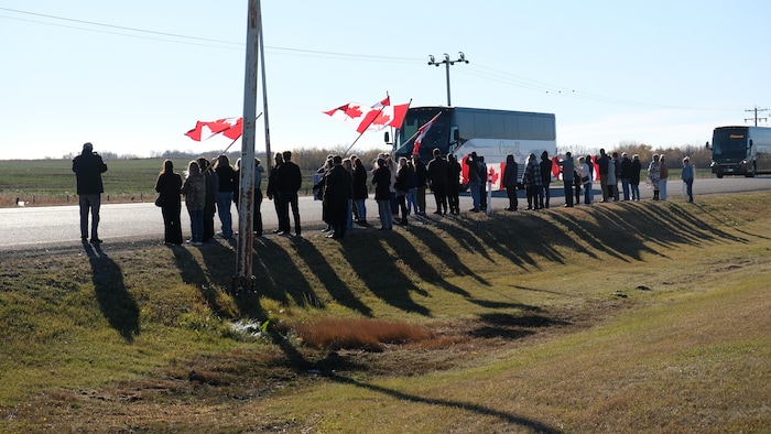 Des personnes rassemblées, certaines avec des drapeaux canadiens, alors que des autobus passent sur l'autoroute à Wainwright le 21 octobre 2026. 