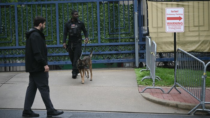 Un policier accompagné d'un chien pisteur se tient devant les grilles d'un immeuble à côté d'un panneau indiquant qu'il s'agit du stationnement de l'ambassade d'Israël.