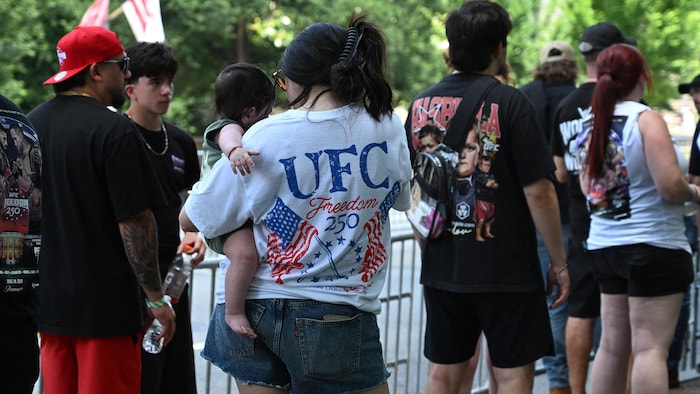 Une femme arborant un t-shirt « UFC » et tenant un bébé dans ses bras fait la file sur le trottoir.