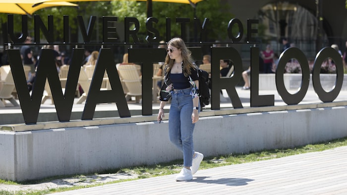 Une étudiante marche sur le campus de l'Université de Waterloo devant une enseigne géante avec le nom de l'université.