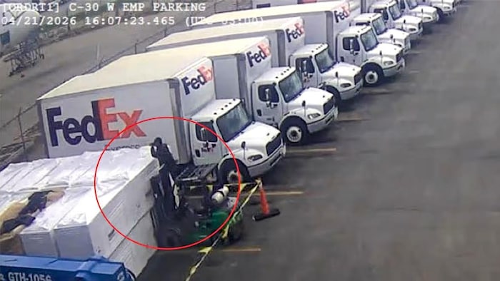 Un homme tient un appareil photo debout sur un chariot élévateur stationné, à côté d’une rangée de camions FedEx.