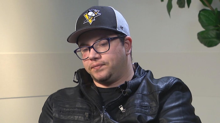 Un jeune homme assis, l'air songeur. Il porte une veste de cuir, des lunettes et une casquette des Penguins de Pittsburgh.