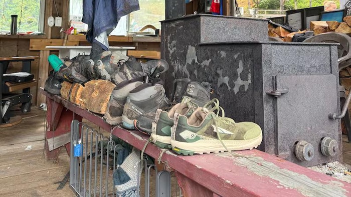 Des chaussures de randonnée mouillées sont alignées sur une planche en bois près d'un poêle.