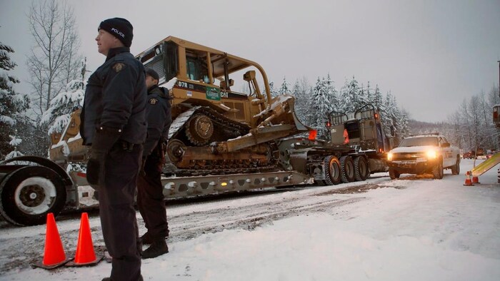 Des policiers, en hiver, se trouvent au bord d'une route dans la forêt. Des machines lourdes sont vues près d'eux.