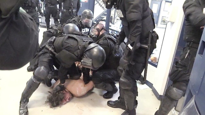 Plusieurs agents des forces de l'ordre en tenue tactique maîtrisant violemment William Ahmo, torse nu au sol, blessé.