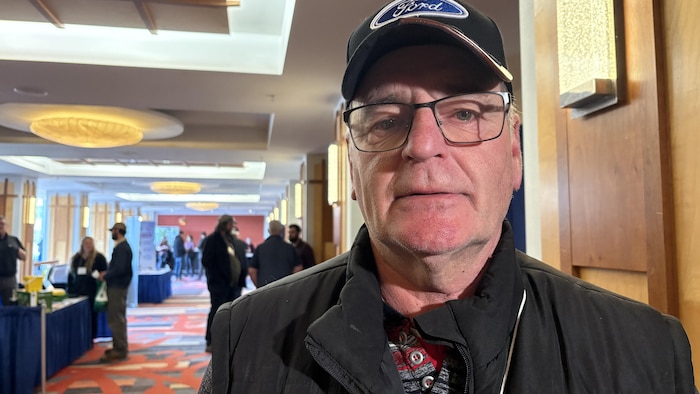 Un homme coiffé d'une casquette décorée du logo de la compagnie Ford est dans le couloir d'un hôtel pour une conférence et regarde droit devant lui.
