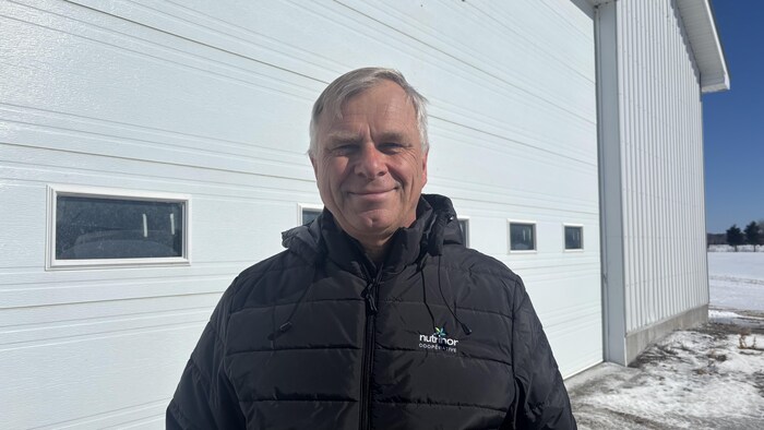 Un homme d'âge mûr sourit devant un grand bâtiment blanc aux portes de garage horizontales. Il porte un manteau noir matelassé identifié au logo de la coopérative Nutrinor. L'arrière-plan montre un paysage enneigé sous un ciel bleu clair.