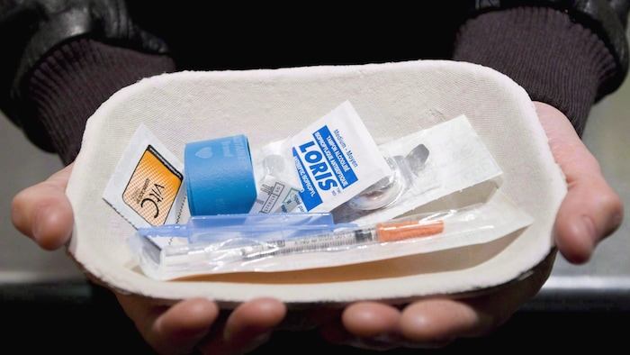 Deux mains tiennent un petit plat de carton avec tous les objects stérilisés nécessaires à l'injections de drogue comme une seringue. 