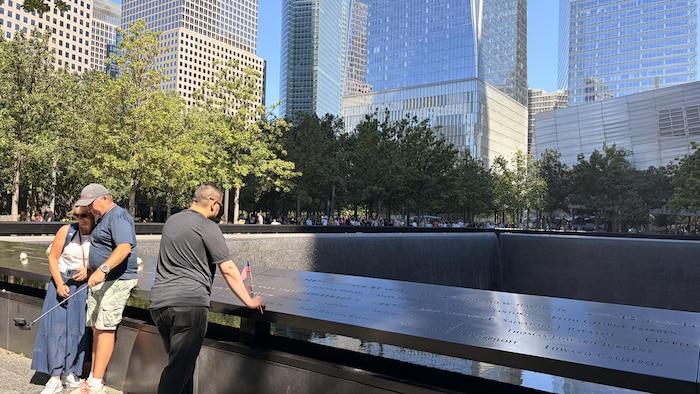 Un homme portant un pantalon et un tshirt noir regarde devant lui à côté d'un couple qui se prend en photo.