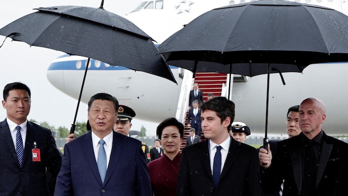 Gabriel Attal et Xi Jinping, côte à côte et suivis de Peng Liyuan, marchent sur un tarmac entourés de gens qui tiennent des parapluies. 