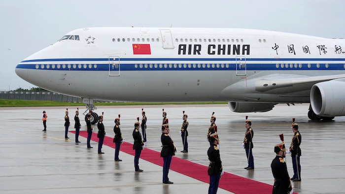 Un avion d'Air China portant le drapeau de la Chine est garé sur le tarmac à côté d'un long tapis rouge bordé d'employés vêtus de costumes protocolaires.