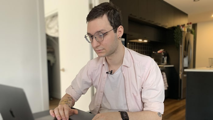 Un jeune homme pose devant la caméra. Il est assis à une table et travaille sur son ordinateur.