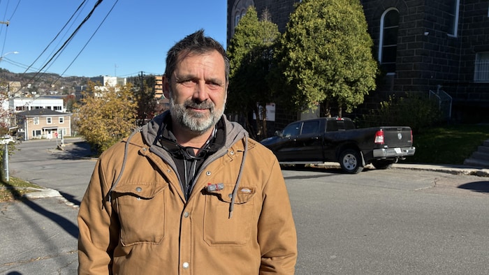 Un homme barbu portant un manteau brun clair se tient à l'extérieur.