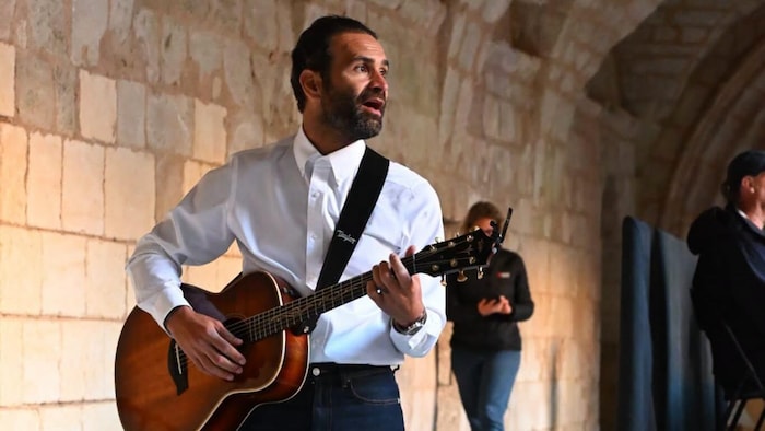 Un homme portant une chemise blanche et un pantalon noir joue de la guitare.