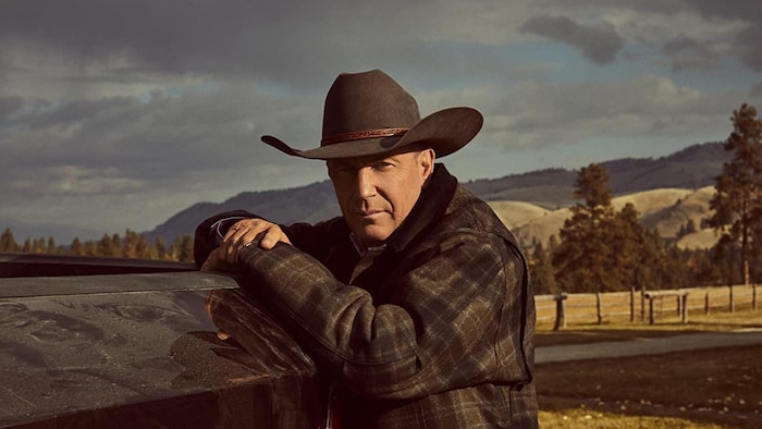L'homme portant une veste à carreaux et un chapeau de cowboy est appuyé sur la caisse découverte d'une camionnette. On aperçoit derrière lui un paysage vallonné. 