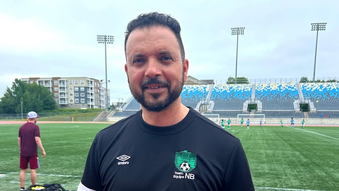Un adulte vêtu d'un chandail de Soccer NB pose dehors pour une photo, debout sur la piste d'athlétisme qui entoure le terrain de football.