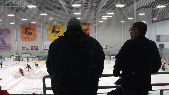 Deux hommes regardent du haut de la patinoire une partie de hockey.
