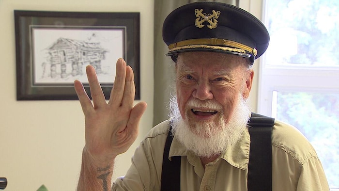 Dick Stevenson portant une casquette de capitaine de bateau sourit en saluant de la main.