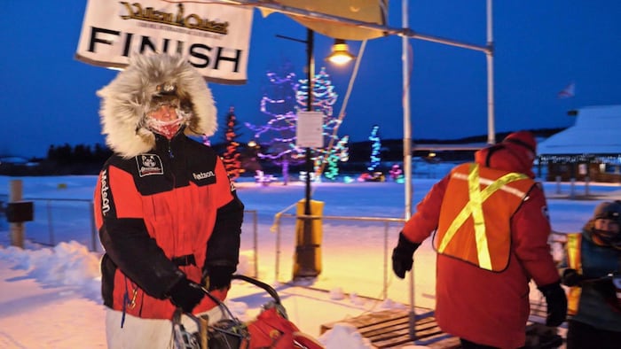 Une femmes sur une traîneau à chiens. Elle est beaucoup habillée. Il y a du gel sur son écharpe. Derrière elle, il y a une banderole sur laquelle il est indiqué : Yukon Quest Finish.
Il fait nuit. À droite, une personne est de dos. Elle porte un gilet fluorescent.