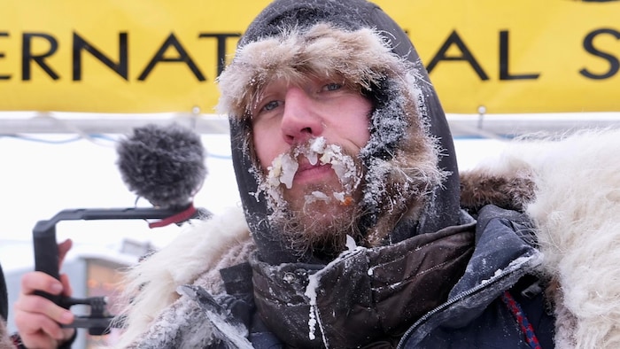 Gros plan sur la tête d'un homme qui porte un gros manteau d'hiver et une capuche. Sa moustache est entièrement gelée.