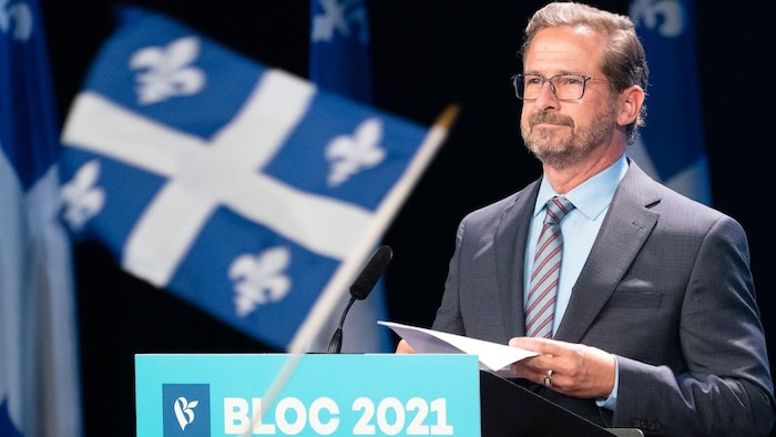 Yves-François Blanchet giving speech in front of microphone with Québec flags in background. Someone is also waving a Québec flag in front of camera.