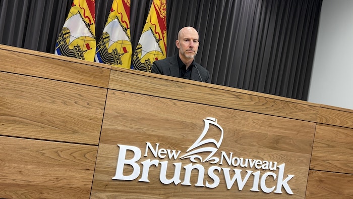 Yves Léger, devant un micro, des drapeaux du Nouveau-Brunswick derrière lui.