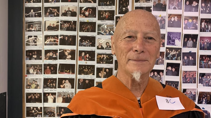 Une personne debout avec une toge universitaire fixe l'appareil photo.