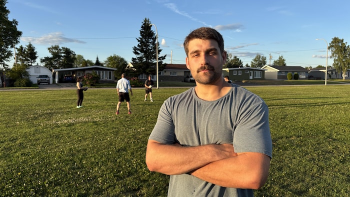 Un homme jeune avec une moustache et les bras croisés se tient debout sur un terrain d'herbe en fin de journée.