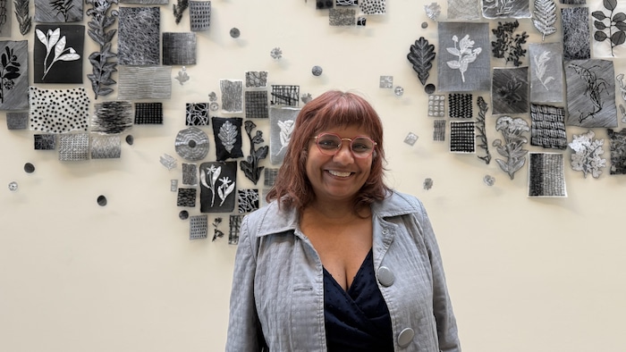 Zahra McDoom, conservatrice des collections à Museum London, sourit devant une œuvre murale composée de nombreux motifs végétaux en noir, blanc et gris.