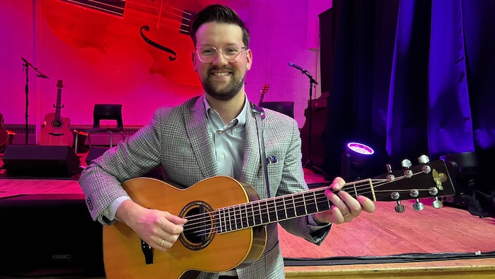 Zak Cormier, en veston gris à carreaux, tient une guitare acoustique et pose pour une photo, en bas de la scène.