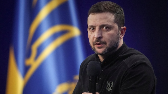 Volodymyr Zelensky parle dans un micro face à un drapeau arborant les armoiries ukrainiennes.
