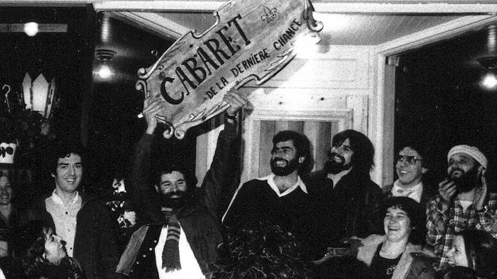 Sept personnes, dont une tenant l'affiche du Cabaret de la dernière chance à bout de bras, devant l'établissement.