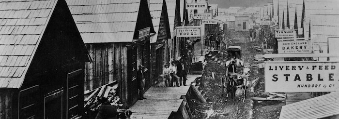 Black and white photo of Barkerville's main street with a carriage in the center and people on the terraces of wooden houses.