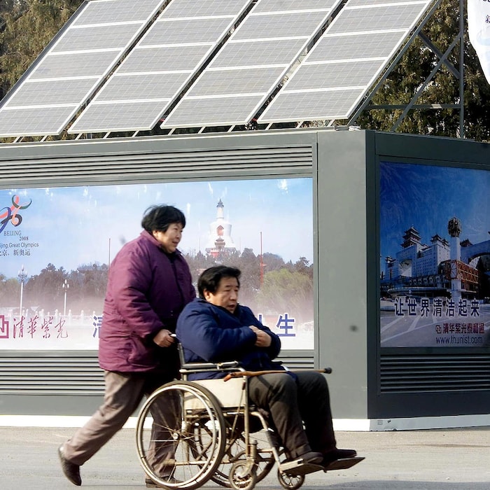 Une femme pousse un homme dans un fauteuil roulant à l'extérieur, devant une publicité surmontée de panneaux solaires
