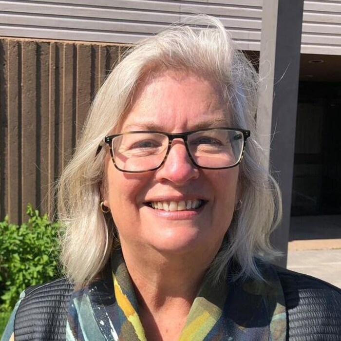 Une femme portant un blouson aux motifs indéfinissables et portant un gros macaron du Parti vert à la boutonnière est debout à l'extérieur devant un bâtiment et sourit en regardant l'objectif de la caméra.