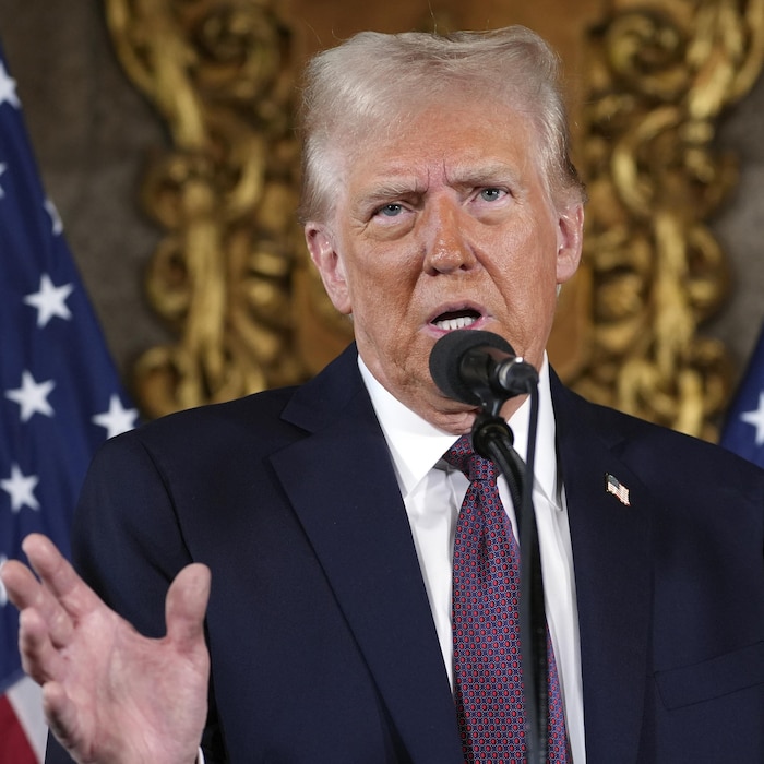 Donald Trump au micro, devant des drapeaux américains, bouge la main en parlant.