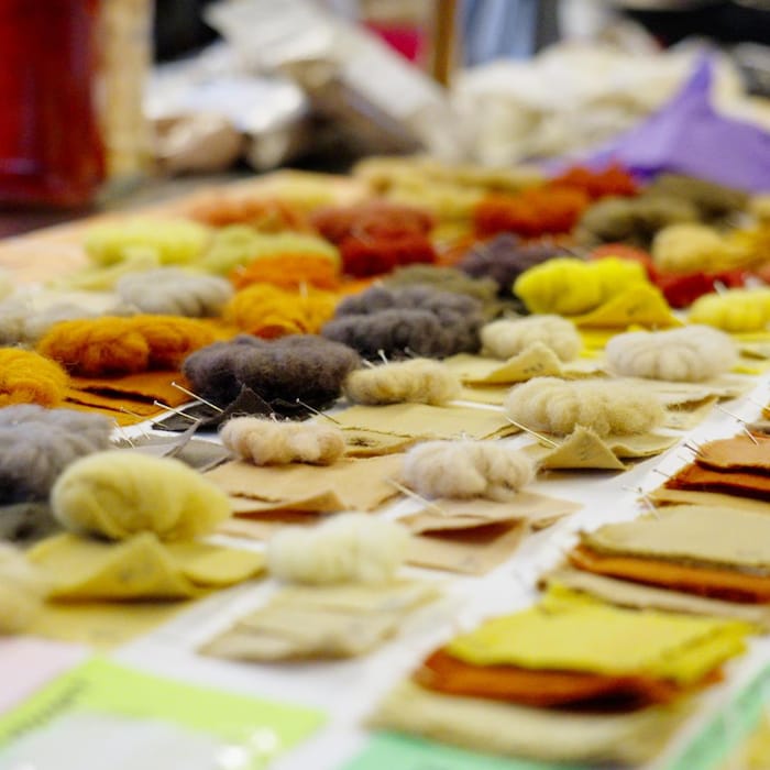 Un plateau sur lequel reposent des carrés de tissus et des pelottes de laines colorés en tons orangés, jaunes, bruns et noirs.