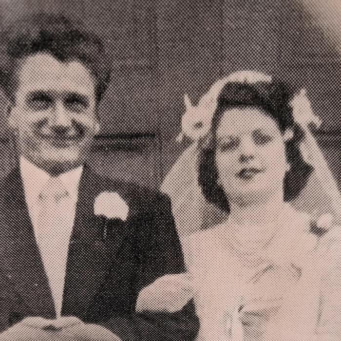 Cette photographie d'époque en noir et blanc présente un couple de mariés. L'homme porte un costume sombre avec une boutonnière et tient un chapeau. La femme porte une robe de mariée claire, un voile orné de fleurs et un bouquet.