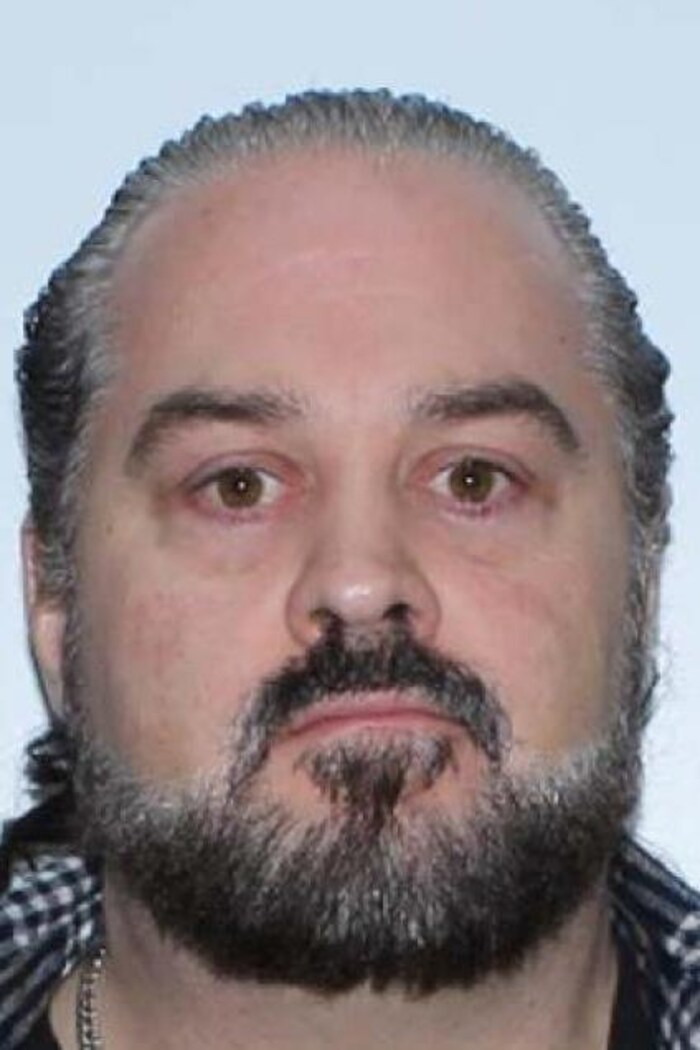 Un homme barbu qui pose devant un mur blanc. 