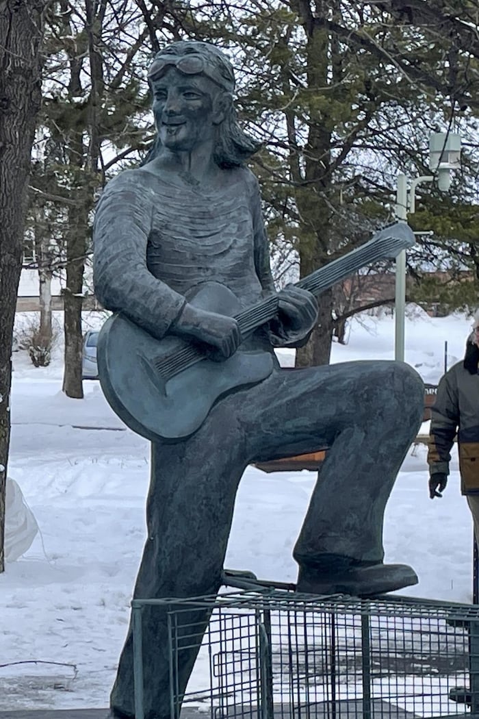 ne statue en bronze d'un homme jouant de la guitare, juché sur un chariot d'épicerie, au centre d'un parc enneigé. À gauche, un petit groupe de personnes est rassemblé sous une tente noire.