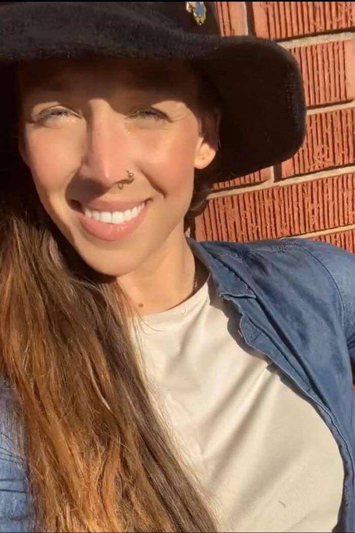 Une femme photographiée au soleil devant un mur de brique. Elle porte un grand chapeau noir et sourit.