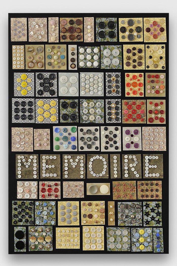 Un panneau, vu de près, où sont cousus des centaines de boutons de vêtement colorés. Des boutons blancs sont placés de manière à ce qu'on puisse lire le mot « mémoire ».