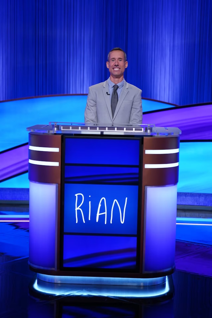 Rian Robertson, vainqueur de « Jeopardy! », se tient debout derrière son pupitre, tout sourire. 