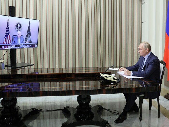 Le président russe, assis au bout d'une longue table, regarde un écran placé à distance sur lequel on voit Joe Biden.