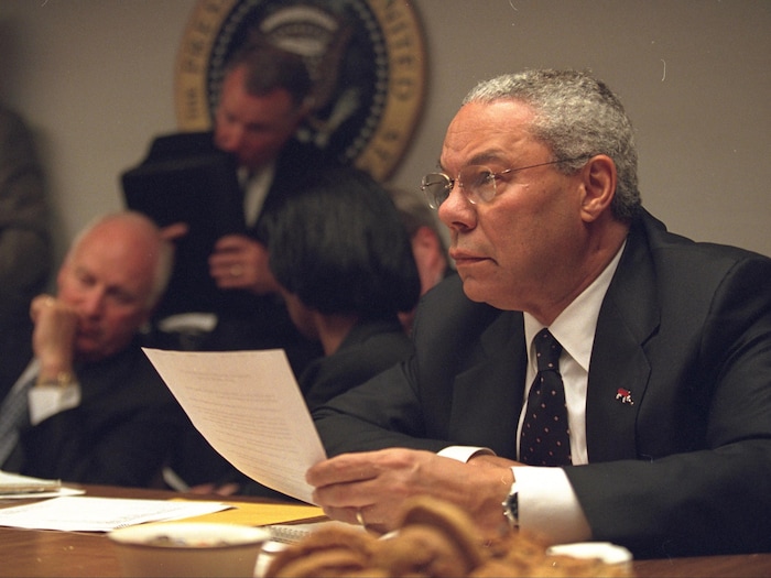 Colin Powel l'air sérieux, un document à la main, assis à une table.