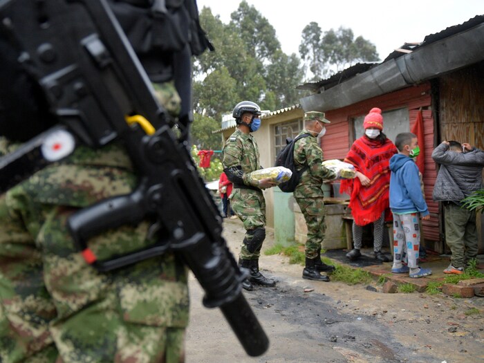 Des soldats armés et masqués remettent un sac de nourriture à une femme devant une maison en tôle.