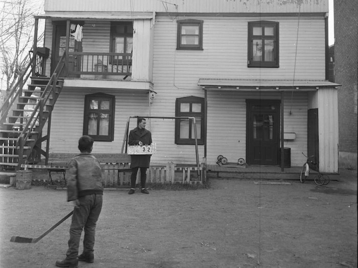 Un jeune garçon tenant un bâton de hockey regarde un homme et son affiche numérotée devant une maison. 