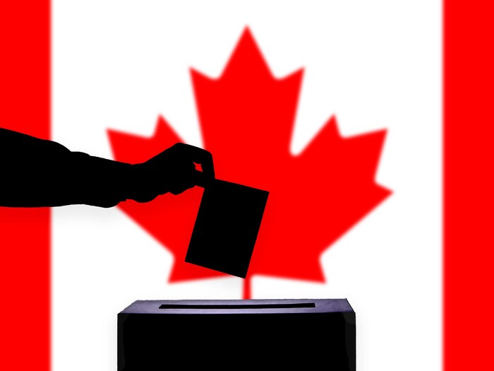 Une personne dépose son bulletin de vote dans une urne devant un drapeau canadien.