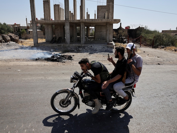 Trois hommes assis sur une moto, dont un qui tient une arme, passent devant une structure d'édifice en béton pendant qu'un nuage de fumée s'élève de débris calcinés sur le bord de la route.