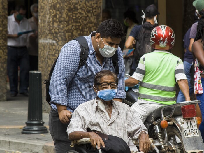 Un homme, le visage couvert d'un masque, pousse un fauteuil roulant en ville.
